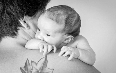 Ein kleines Baby schaut neugierig über die Schulter einer erwachsenen Person, deren Tattoo eines Efeublattes auf der Schulter sichtbar ist. Das Schwarz-Weiß-Foto fängt einen zärtlichen und geschützten Moment ein. Eine liebevolle Aufnahme von Fotograf Michael Schalansky.